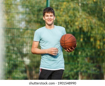 A Cute Young Boy Plays Basketball On The Street Playground In Summer. Teenager In A Green T-shirt With Orange Basketball Ball Outside. Hobby, Active Lifestyle, Sports Activity For Kids.