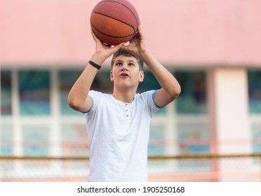 A cute young boy plays basketball on the street playground in summer. Teenager in a white t-shirt with orange basketball ball outside. Hobby, active lifestyle, sports activity for kids. - Powered by Shutterstock