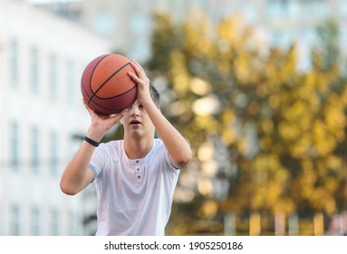 A Cute Young Boy Plays Basketball On The Street Playground In Summer. Teenager In A White T-shirt With Orange Basketball Ball Outside. Hobby, Active Lifestyle, Sports Activity For Kids.