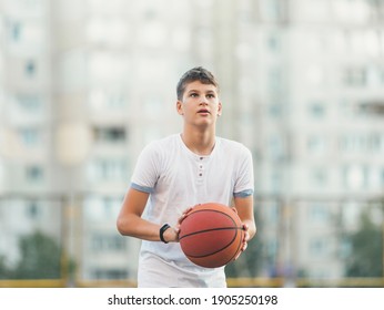 A Cute Young Boy Plays Basketball On The Street Playground In Summer. Teenager In A White T-shirt With Orange Basketball Ball Outside. Hobby, Active Lifestyle, Sports Activity For Kids.