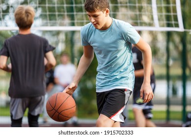 A cute young boy plays basketball on the street playground in summer. Teenager in a green t-shirt with orange basketball ball outside. Hobby, active lifestyle, sports activity for kids.