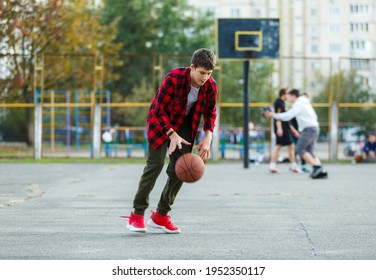 Cute Young Boy Plays Basketball On Street Playground. Teenager In Red Flannel Checked Shirt With Orange Basketball Ball Outside. Hobby, Active Lifestyle, Sport Activity For Kids.