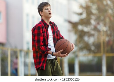 Cute Young Boy Plays Basketball On Street Playground. Teenager In Red Flannel Checked Shirt With Orange Basketball Ball Outside. Hobby, Active Lifestyle, Sport Activity For Kids.