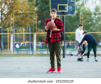 Cute Young Boy Plays Basketball On Street Playground. Teenager In Red Flannel Checked Shirt With Orange Basketball Ball Outside. Hobby, Active Lifestyle, Sport Activity For Kids.