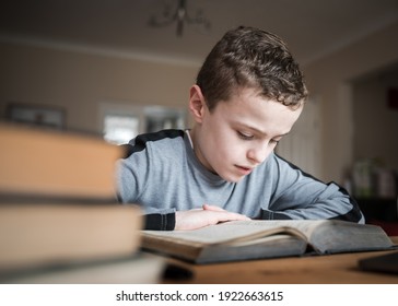 Cute young boy sat reading big old book concentrating hard at wooden table homeschool handsome and focused intensely on learning in front room with pile of books next to him