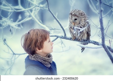 A cute young boy in a warm knitted clothes looking at the owl sitting on the branch covered with frost on a frosty winter day. Kids and nature.