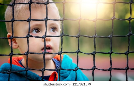 Cute Young Caucasian Boy Looking Through The Tennis Net In Spring