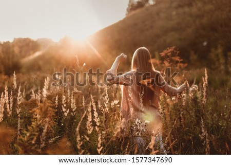 Cute young caucasian girl with long fluffy hair in a field at sunset. Beautiful young woman with brown hair dancing in a field at sunset. Freedom and nature concept.Soft focus