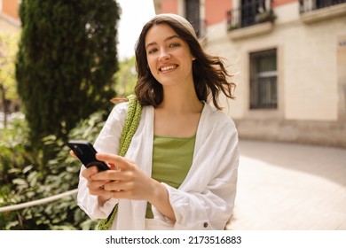 Cute Young Caucasian Girl With Phone Looks At Camera Standing Outdoors. Brunette Wears Bandana, Shirt With T-shirt. Sincere Emotions Lifestyle Concept