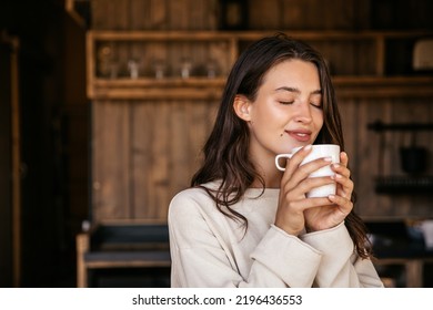 Cute young caucasian woman with closed eyes enjoys fragrant coffee in morning indoors. Brunette wears white sweatshirt. Lifestyle, different emotions, leisure concept. - Powered by Shutterstock