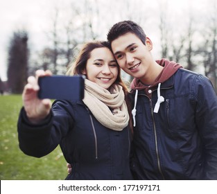 Cute Young Couple Looking Happy While Taking Pictures Using A Smart Phone At The Park. Mixed Race Teenage Boy And Girl In Love Photographing Themselves Outdoors.