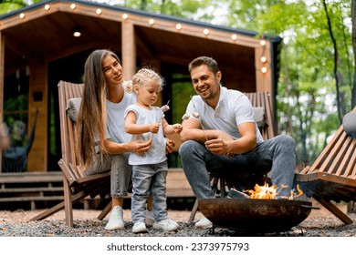 Cute young family dad mom and daughter are sitting together by a fire in the forest and roasting marshmallows near their country house - Powered by Shutterstock