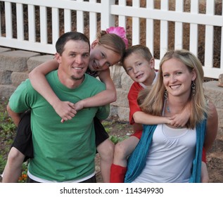 A Cute Young Family Doing Piggy Back Rides In Their Picket Fenced Back Yard.