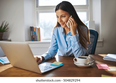 Cute Young Female Adult With Grin On Phone While Working On Laptop Computer At Desk Next To Coffee Cup With Large Window In Background