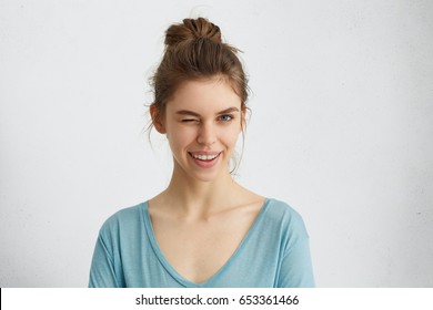 Cute young female with delicate features having hair tied in knot wearing blue sweater blinking her eyes with pleasure having happy expression. Facial expressions and people emotions concept