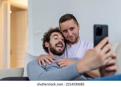 Cute Young Gay Couple Video Calling Their Friends In Their Living Room At Home. Two Male Lovers Smiling Cheerfully While Greeting Their Friends On A Smartphone. Young Gay Couple Sitting Together.