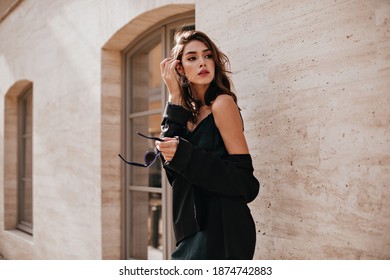 Cute young girl with dark wavy hairstyle and bright makeup, silk dress, black jacket, holding sunglasses in hands and looking away against beige building background - Powered by Shutterstock