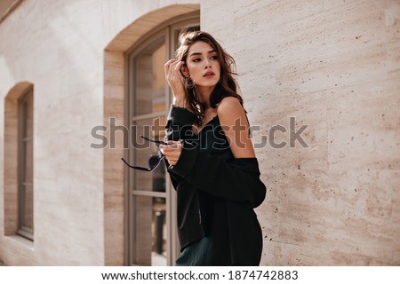 Cute young girl with dark wavy hairstyle and bright makeup, silk dress, black jacket, holding sunglasses in hands and looking away against beige building background