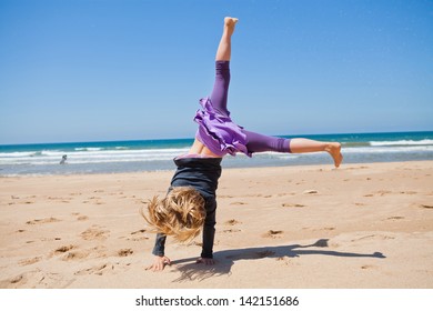 Cute Young Girl Doing Cartwheel In Sand At Beach With Blue Sky And Ocean In Background