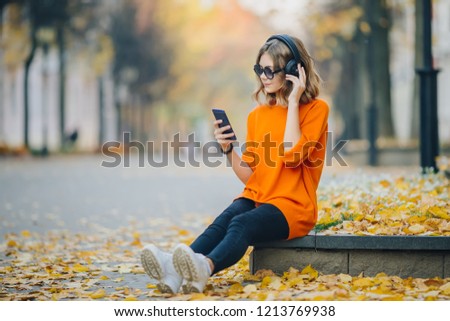 cute young girl listening music in headphones, urban style, stylish hipster teen sitting on a sidewalk on city street and choosing track on mobilephone infront of oidsity buildings, orange street