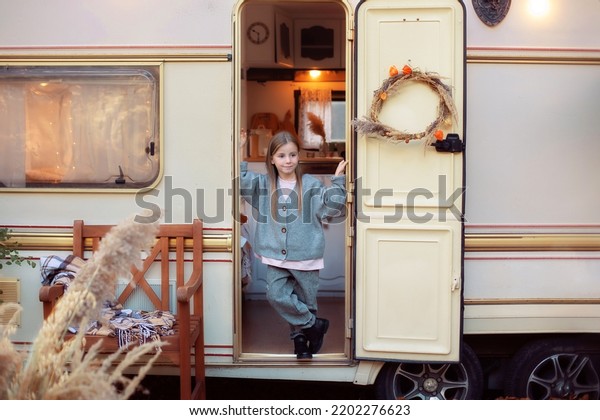 Cute young girl stand near trailer door. Child in cozy\
campsite fall backyard. Concept camping, outdoor, nature,\
adventure. Smiling little girl in casual clothes standing on porch\
RV house in garden. 