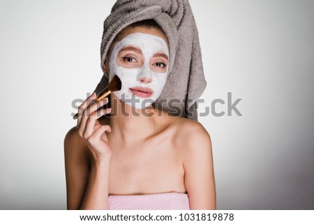 cute young girl with a towel on her head puts a white moisturizing mask on her face, using a brush