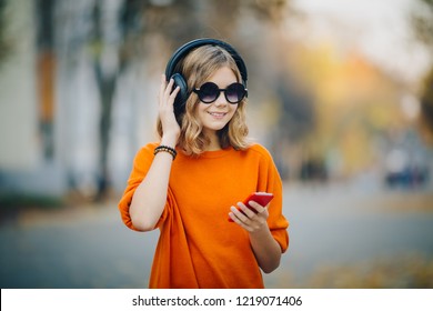 cute young girl walking down old city street and listening music in headphones, urban style, stylish hipster teen hold mobile phone and and smile, orange crazy style