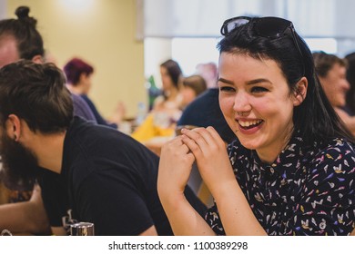 Cute Young Hip Woman With Black Hair, Septum Piercing And A Big Smile Sitting In A Restaurant And Smiling Among Other People.