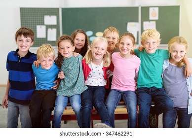 Cute Young Learners Inside Their Classroom, Smiling At The Camera While Holding Each Other By Shoulder.