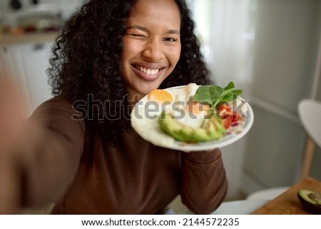 Cute young lovely adorable female of black ethnicity doing selfie over kitchen background holding plate with colorful healthy and nutritious breakfast of eggs and vegetables, winking at camera