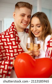 Cute young man and beautiful girl with long brown hair are standing gently embracing under one blanket . They celebrate Valentine's Day with glasses of wine and red balloon heart. Love and romance. 
