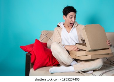 A Cute Young Man In Pajamas Sits On The Couch With A Box Of Pizza. Beige Sofa And Red Pillows. Rest In The Evening With Food. Student Life. Blue Background.