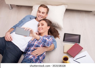 Cute Young Married Couple Is Making Selfie At Home. They Are Lying On Flooring And Smiling. The Woman Is Holding A Mobile Phone. They Are Embracing