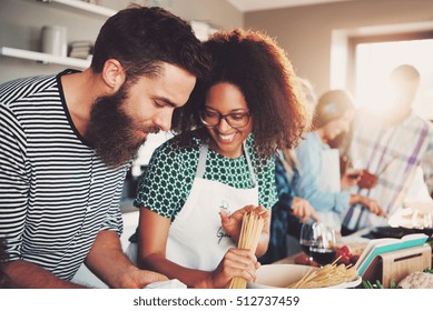 Cute Young Mixed Couple Preparing Pasta Dinner In Kitchen With Friends. Bright Sunlight Shining Through Window.