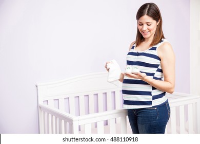 Cute Young Pregnant Woman Holding A Pair Of Baby Shoes And A Onesie Next To A Crib And Smiling