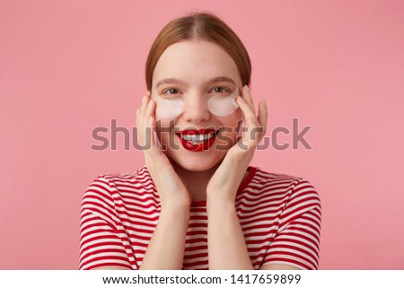 Cute young red-haired woman in a red striped T-shirt, with red lips , touches his face with fingers, very pleased with my new patches from dark circles under my eyes. Stands over pink background.