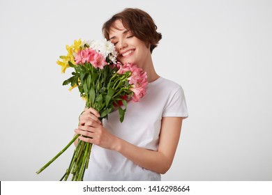 Cute young short haired girl in white blank t-shirt, holding a bouquet of colorful flowers, enjoying the smell, broadly smiling with closed eyes, standing over white background.