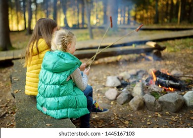 Cute Young Sisters Roasting Hotdogs On Sticks At Bonfire. Children Having Fun At Camp Fire. Camping With Kids In Fall Forest. Family Leisure With Kids At Autumn.