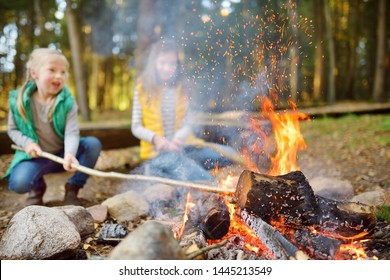 Cute Young Sisters Roasting Hotdogs On Sticks At Bonfire. Children Having Fun At Camp Fire. Camping With Kids In Fall Forest. Family Leisure With Kids At Autumn.