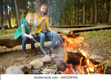 Cute Young Sisters Roasting Hotdogs On Sticks At Bonfire. Children Having Fun At Camp Fire. Camping With Kids In Fall Forest. Family Leisure With Kids At Autumn.
