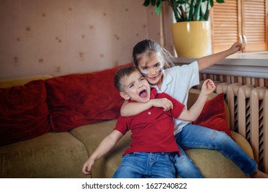 Cute Young Sport Fans, Boy And Girl With Colored Cheeks, Sitting On Sofa, Family Watching Sport Games And Cheering For Her Team On TV At Home, Real Emotions, Sports Event And Fun Supporting Concept