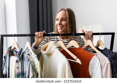 Cute young woman in bathrobe standing in front of hanger rack and trying to choose outfit dressing for work or walk. Selection of a wardrobe, stylist, shopping.