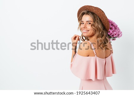 Cute young woman with bouquet fresh flowers wearing straw hat and pink dress smiling looking at camera while standing on white studio background with copy space.