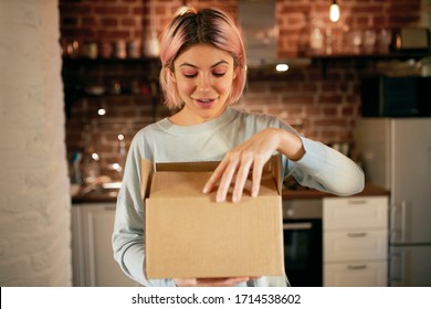 Cute Young Woman In Casual Clothes Standing In Kitchen Interior With Postal Parcel, Unpacking Cardboard Box Full Of Groceries, Having Excited Look. Home Delivery, Purchase And Online Shopping Concept