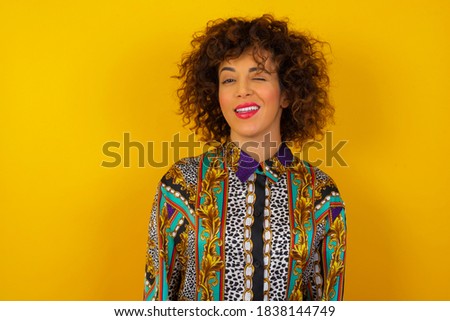 Cute young  woman with curly hair wearing colorful shirt over yellow background blinking her eyes with pleasure having happy expression. Facial expressions and people emotions concept.