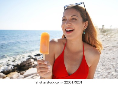 Cute young woman eating an orange popsicle on the beach on summer