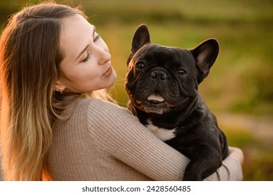 Cute young woman holding French bulldog, smiling at sunset while walking. Favorite dog in the arms of the owner in the park. Concept of pet care, adoption - Powered by Shutterstock