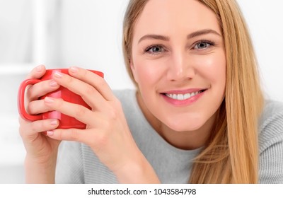 A cute young woman is holding a red cup sitting at a table in a white office. Coffee break - smiling blonde, close-up portrait.