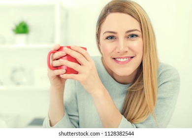 A cute young woman is holding a red cup sitting at a table in a white office. Toned photo.