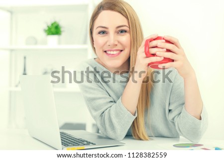 A cute young woman is holding a red cup sitting at a table in a white office. Toned photo.